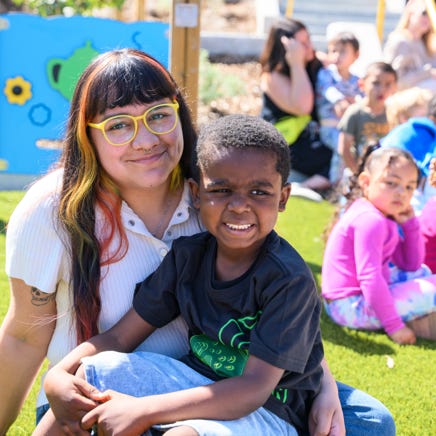 A teacher smiles at the camera while sitting with a young child on their lap.